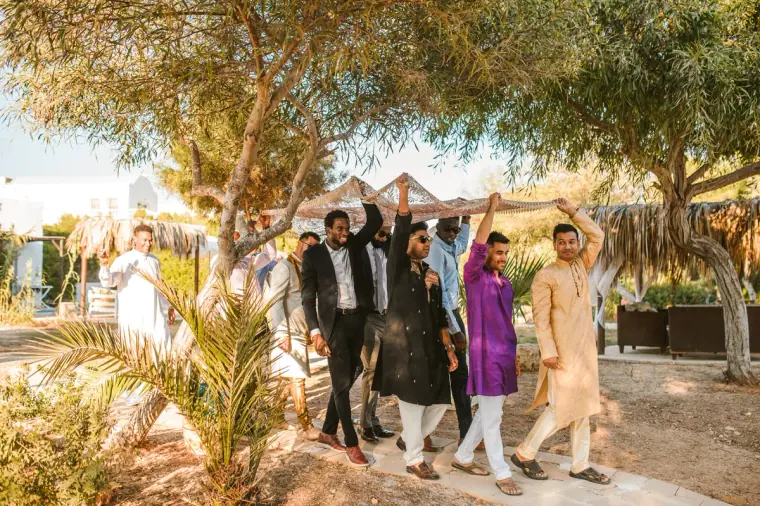 Men in kurtas and suits escorting the groom beneath a canopy along a sandy path
