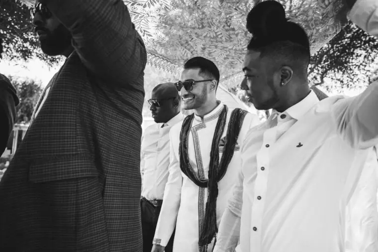 Black-and-white photo of the groom in sunglasses walking beneath the canopy with friends