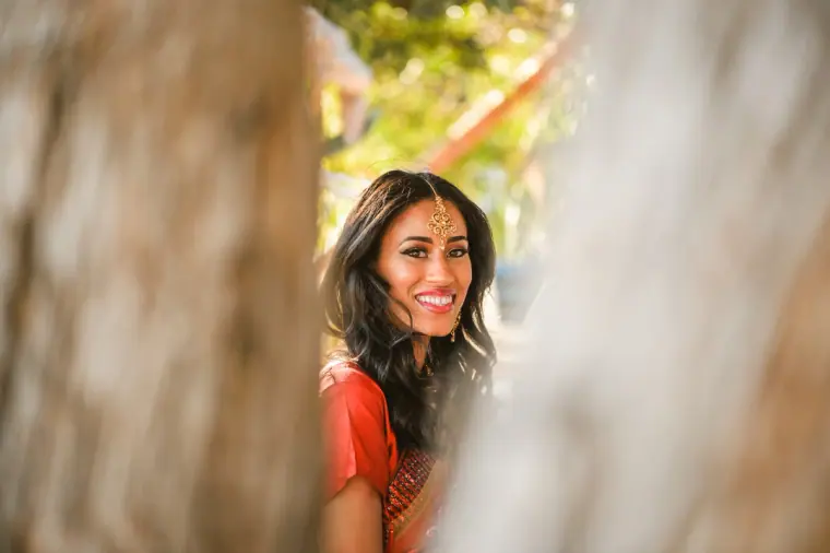Bride in her red sari smiling, glimpsed between two tree trunks