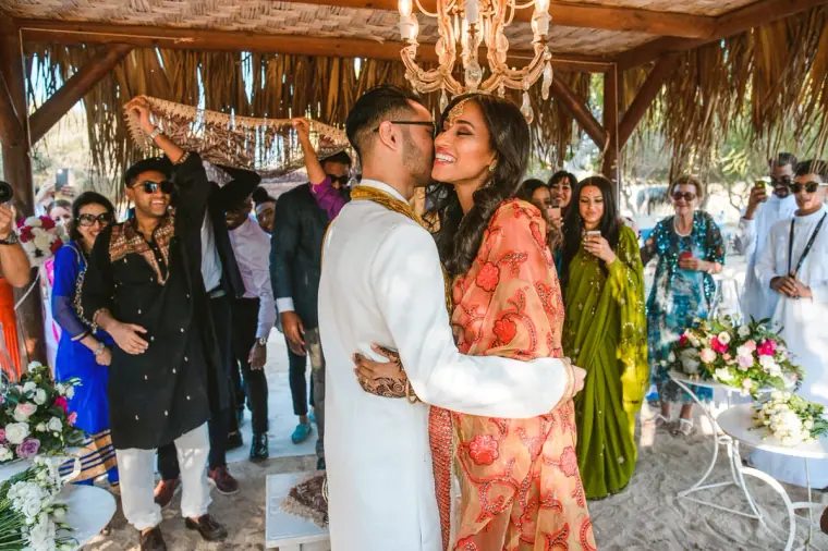 Raya and Adhib embracing beneath the thatched canopy surrounded by guests