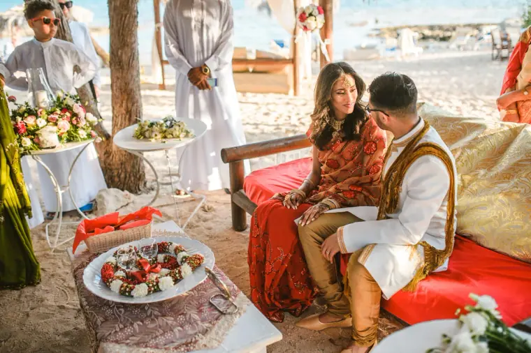 Bride and groom seated on red cushions under the canopy during the traditional ceremony