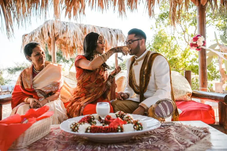 Bride feeding the groom a sweet beside an older relative under the beach canopy