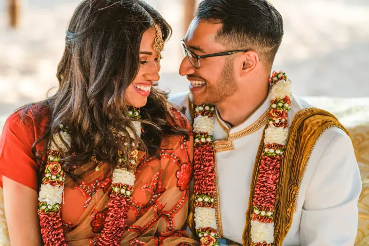 Raya and Adhib in red and white garlands smiling at each other by the beach