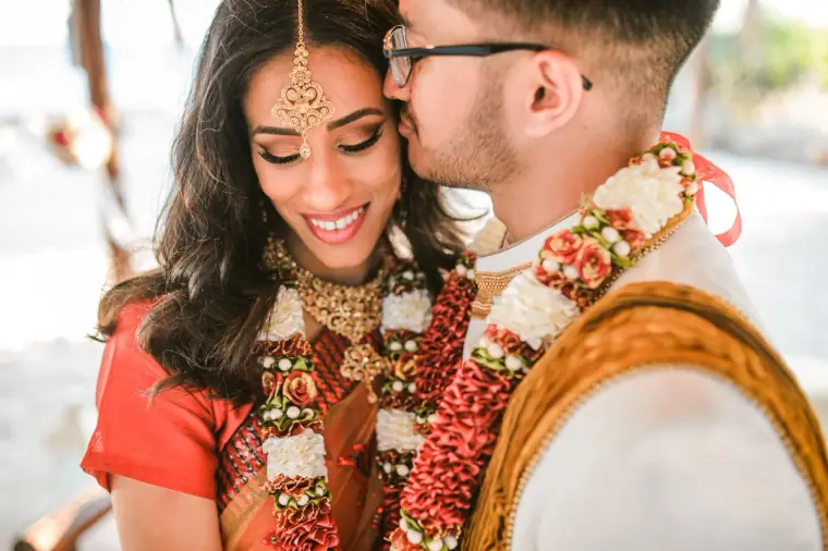 Groom kissing the smiling bride's cheek, both wearing flower garlands