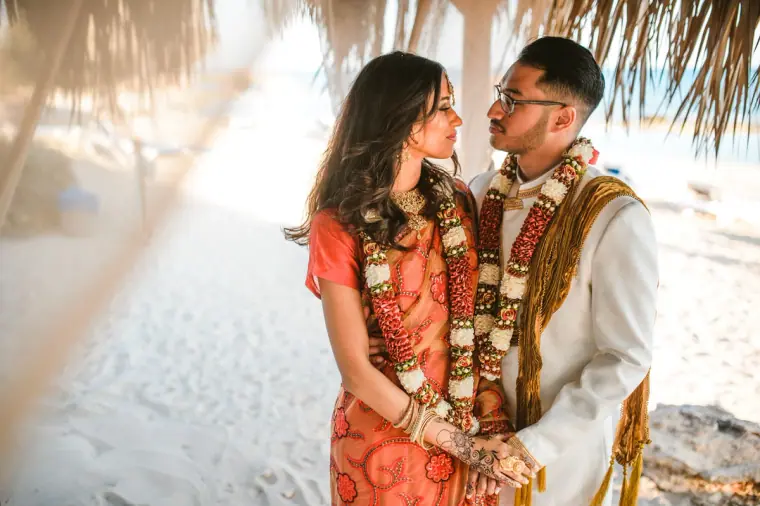 Raya and Adhib in flower garlands facing each other under a draped beach cabana