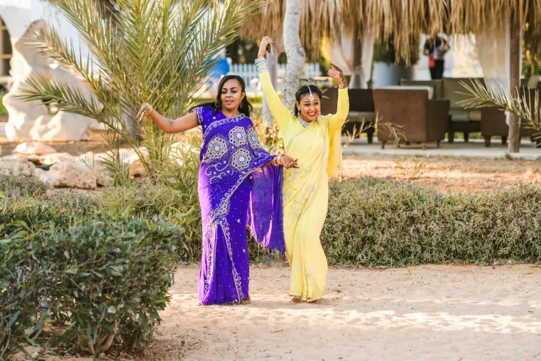 Two women in purple and yellow saris dancing along a garden path by the beach