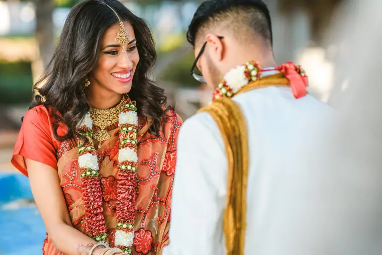 Bride smiling at the groom, both wearing red and white flower garlands