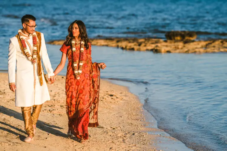 Bride in a red sari and groom in a cream sherwani strolling along the water's edge