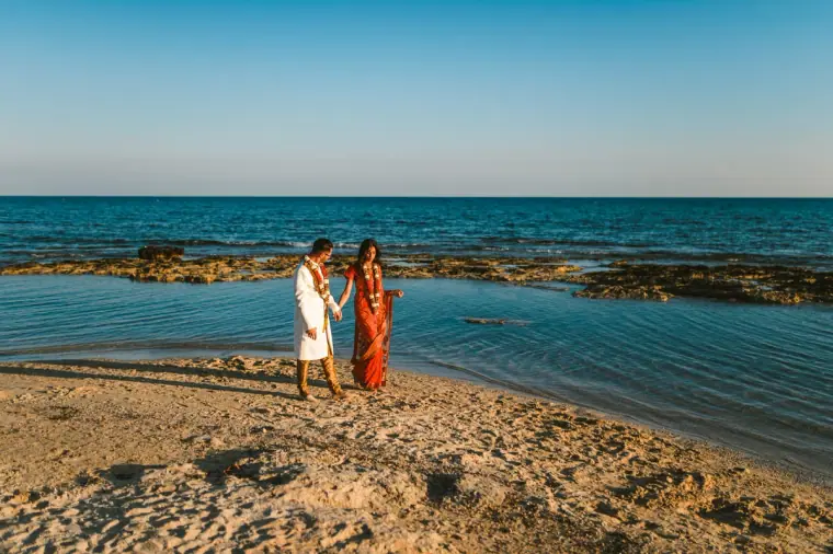 Newlyweds standing on a sandy spit surrounded by calm blue sea