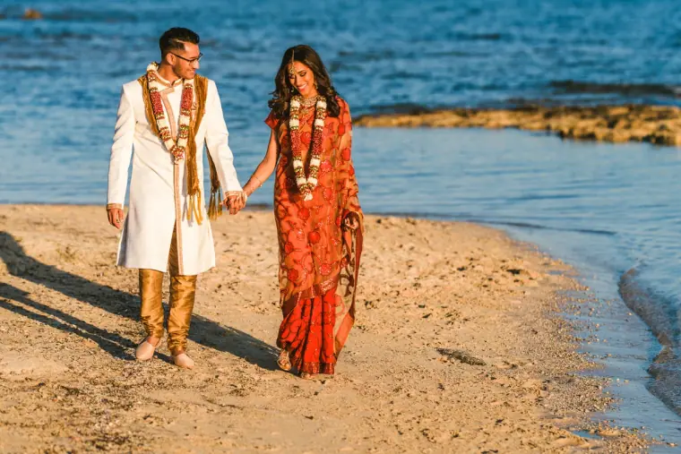 Raya and Adhib walking hand in hand on the beach in their wedding garlands