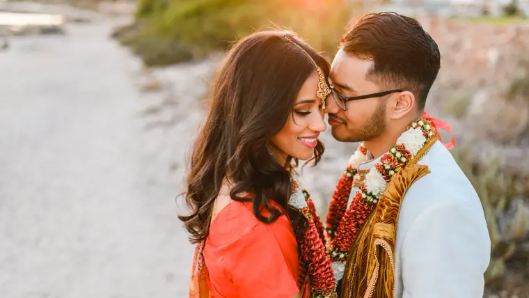 Groom kissing the bride's cheek on the beach path, both in flower garlands