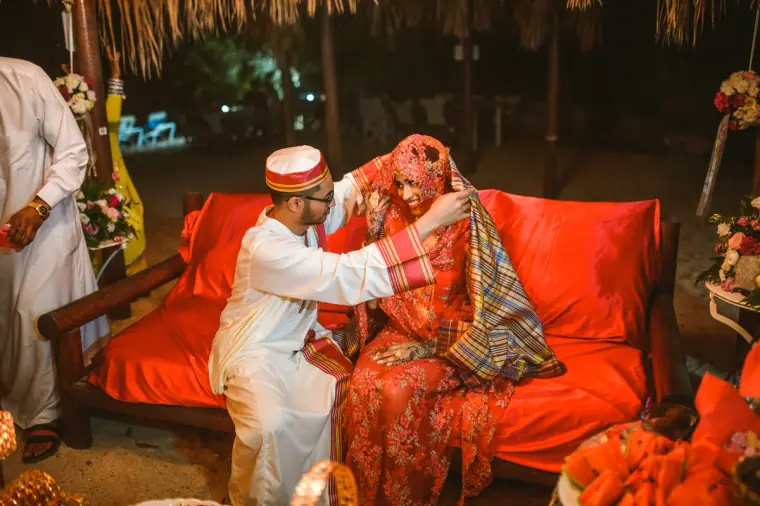 Groom in a white robe and red cap lifting a tartan shawl from the bride's head