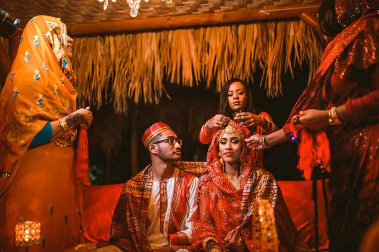 Women gathered around the couple seated on the red daybed under the canopy at night