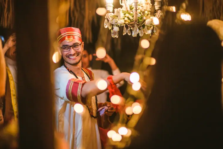 Groom in a red cap smiling beneath the chandelier, framed by fairy lights