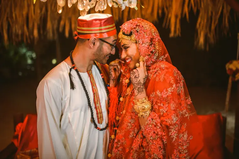 Adhib in a red cap leaning his head against Raya's as she smiles beneath a red veil