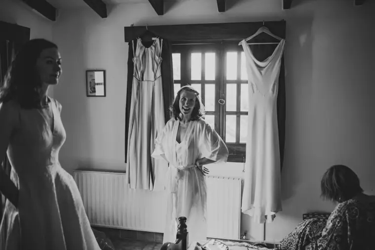 Black-and-white photo of the bride in a robe beside her hanging dress in a beamed room