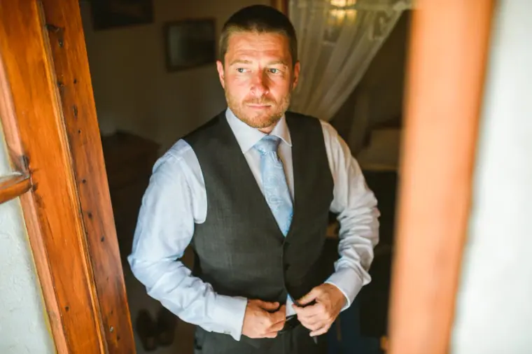 Groom in a grey waistcoat and pale blue tie buttoning up in a doorway
