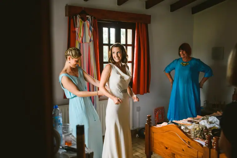Bridesmaid in pale blue fastening the bride's dress in a rustic bedroom