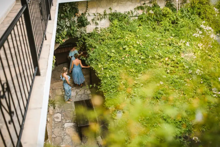 Bride and bridesmaids in blue walking through a leafy stone courtyard, seen from a balcony