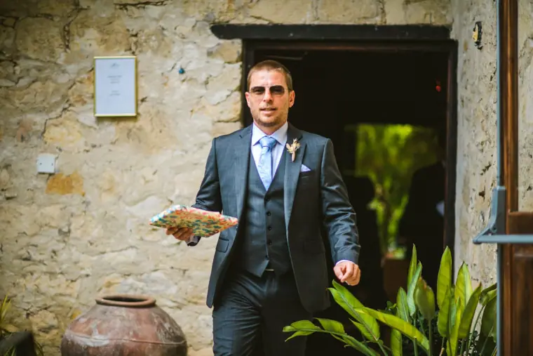 Groom in a grey three-piece suit stepping out of a stone doorway holding a programme