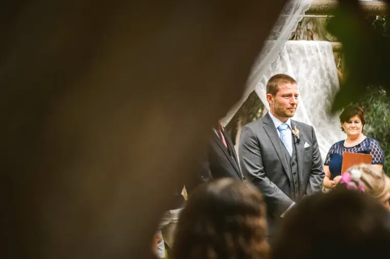 Groom watching the aisle as the bride approaches, glimpsed past guests