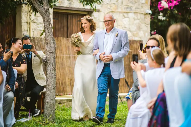Bride walking down the grass aisle on her father's arm as guests take photos