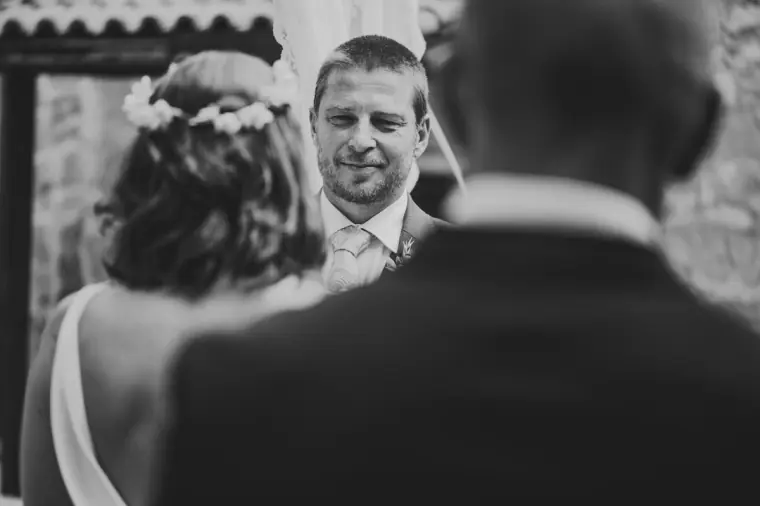 Black-and-white photo of the groom smiling over the officiant's shoulder at the bride