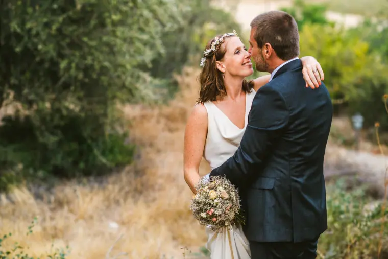 Bride laughing as the groom kisses her cheek, holding a dried-flower bouquet
