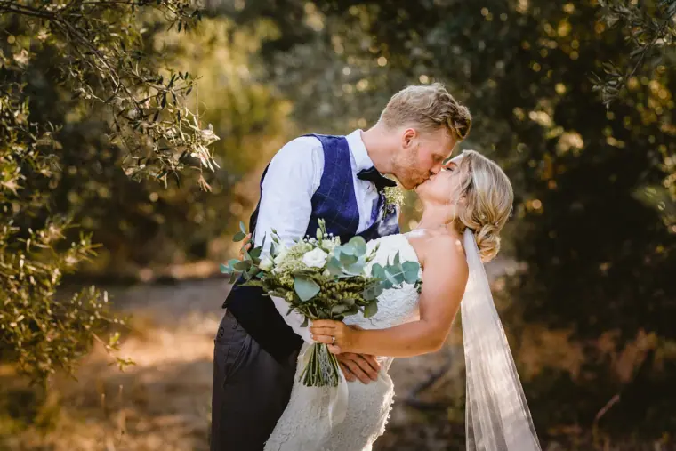 Groom dipping the bride for a kiss in dappled shade, her bouquet in hand