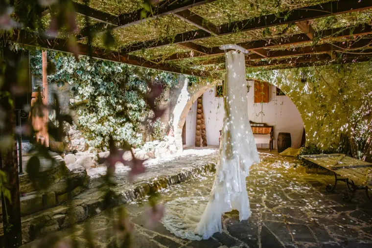 Lace wedding dress hanging from a vine-covered pergola above a stone courtyard
