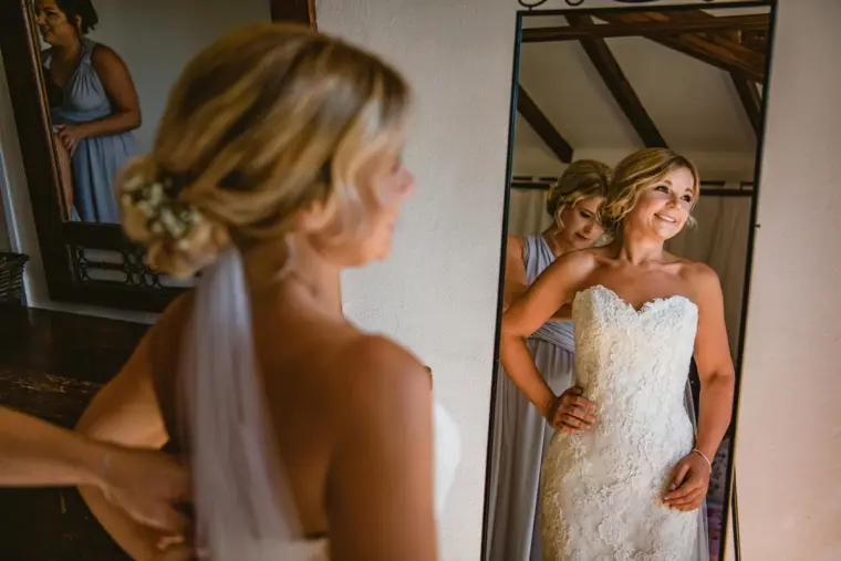 Bride in a lace strapless gown smiling into a mirror as a bridesmaid helps her