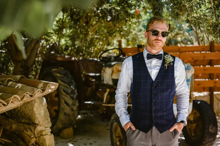 Groom in a navy check waistcoat, bow tie and sunglasses by an old tractor