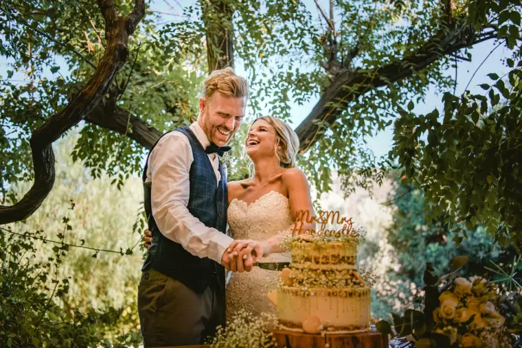 Fiona and Paul laughing as they cut their tiered wedding cake under the trees