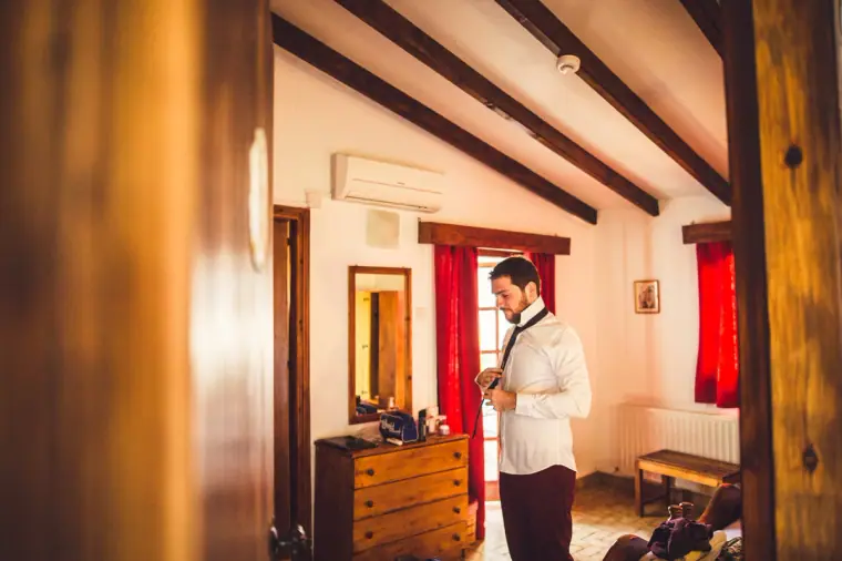 Groom in a white shirt getting ready in a beamed bedroom with red curtains
