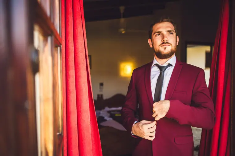 Itai buttoning his burgundy suit jacket beside red curtains