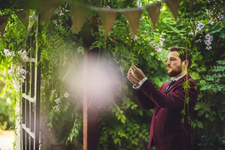 Groom in burgundy adjusting greenery by the ceremony gate, framed by blurred pink blossom