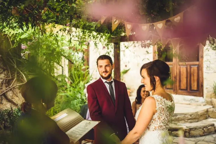 Groom in burgundy and bride in lace standing before the celebrant under the pergola