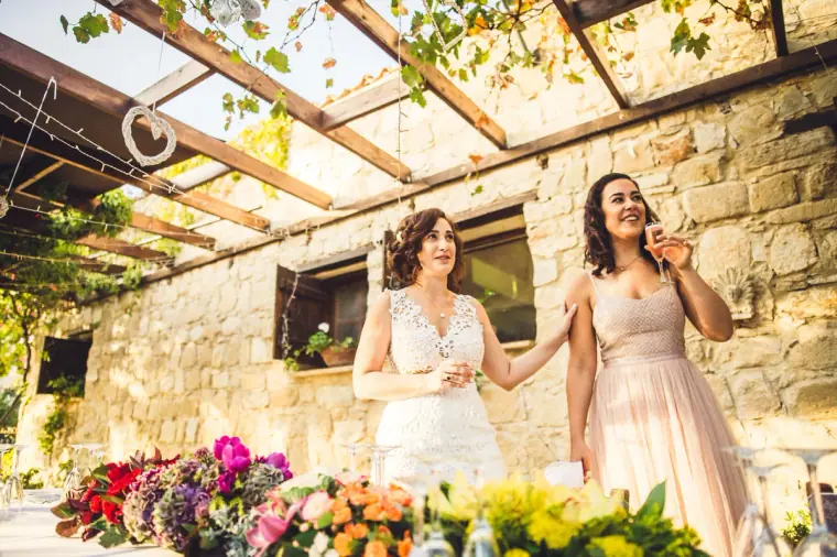 Bride and a friend in blush pink laughing by a long flower-lined table on the terrace