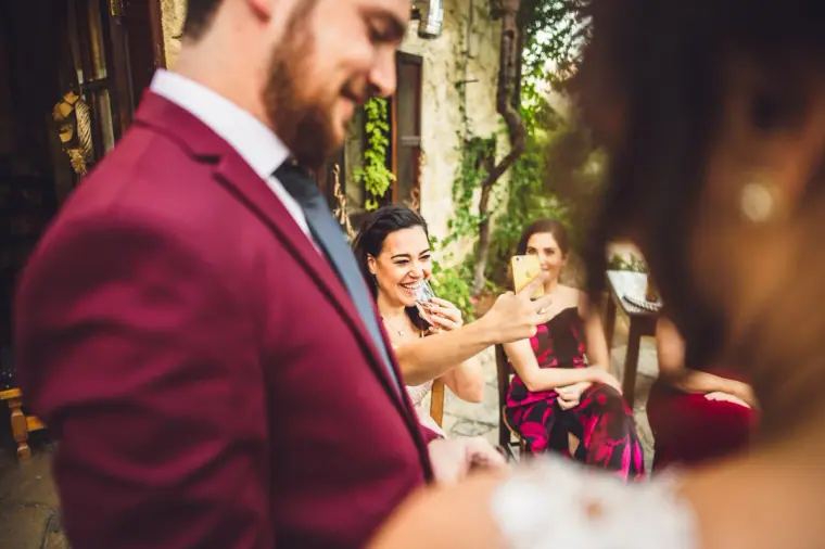 Groom in burgundy laughing as guests take selfies behind him