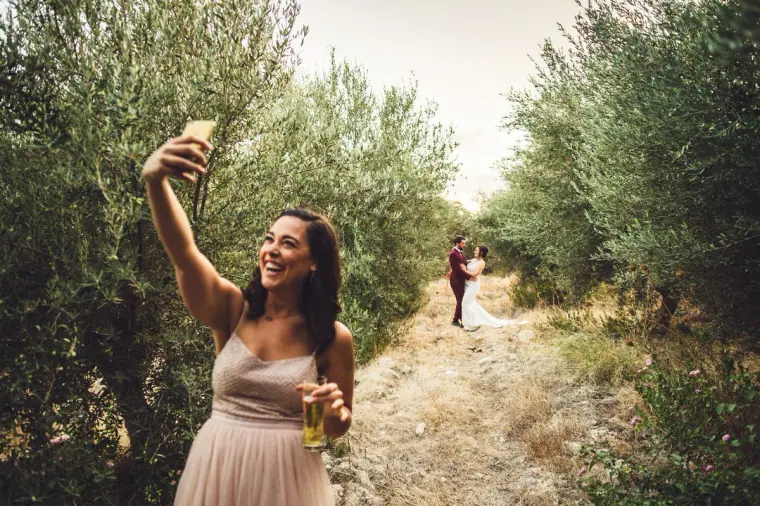 Guest in blush pink taking a selfie as the couple stand far down the olive grove path