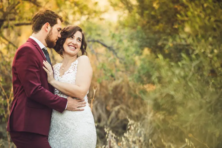 Groom kissing the bride's cheek as she smiles among sunlit trees