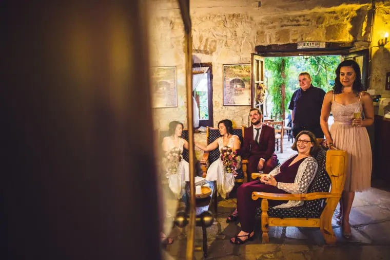 Newlyweds relaxing with guests in the rustic lounge of Vasilias Nikoklis Inn, seen through a doorway