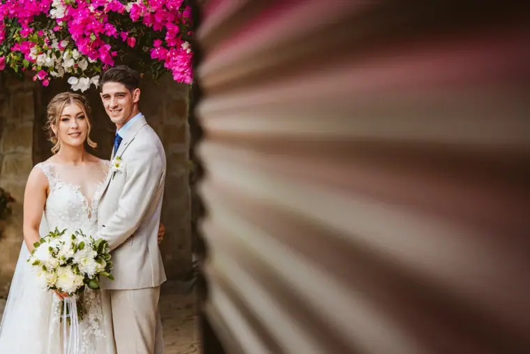 Bride holding a white bouquet beside the groom by pink bougainvillea
