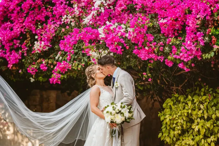 Bride and groom kissing beneath pink bougainvillea, her long veil sweeping out