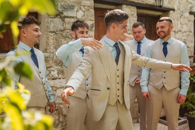 Groom in a beige three-piece suit joking with his groomsmen in the courtyard