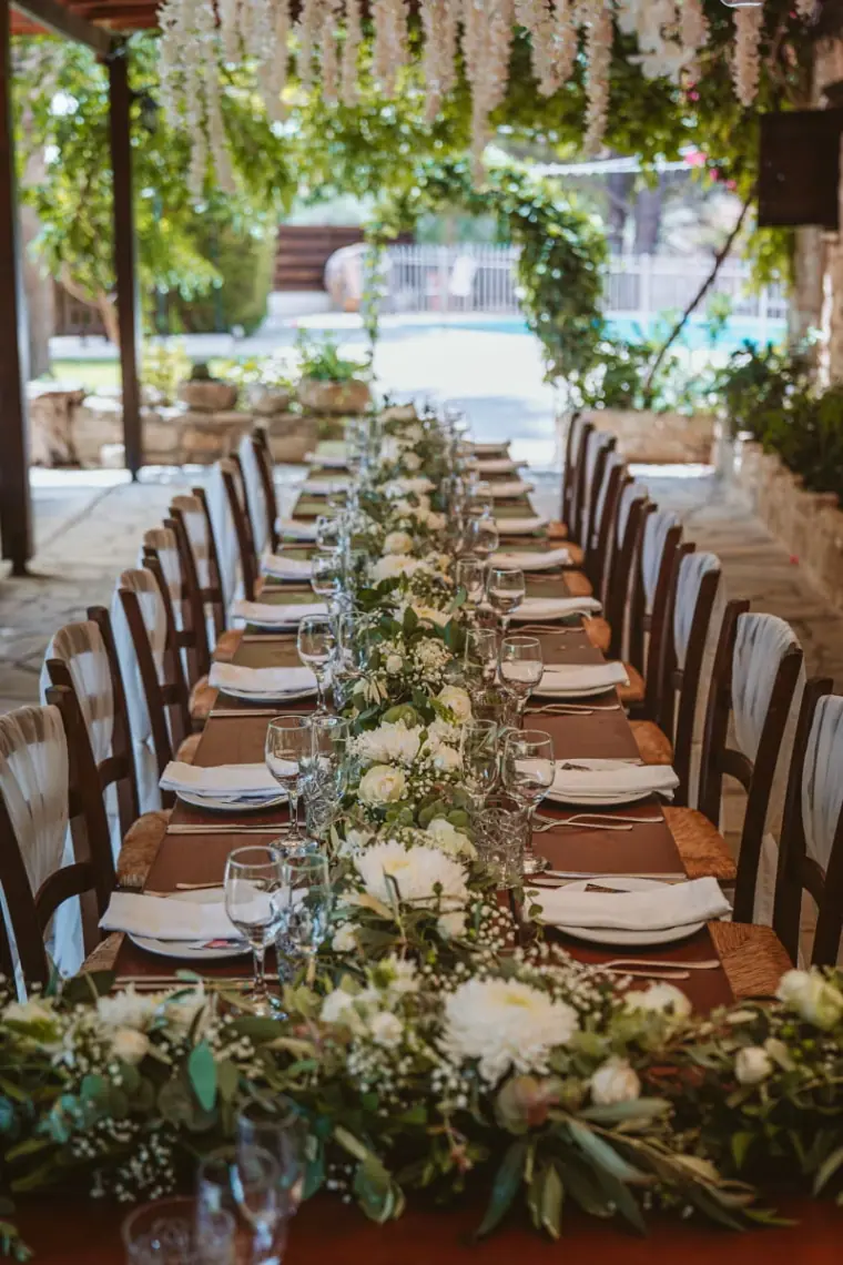 Long rustic banquet table dressed with greenery and white place settings under the pergola