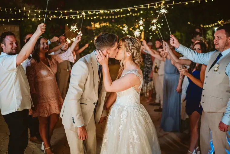 Newlyweds kissing as guests hold sparklers overhead under festoon lights