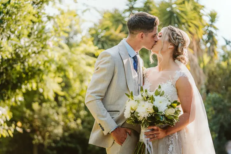 Couple kissing among sunlit trees, white rose bouquet in hand