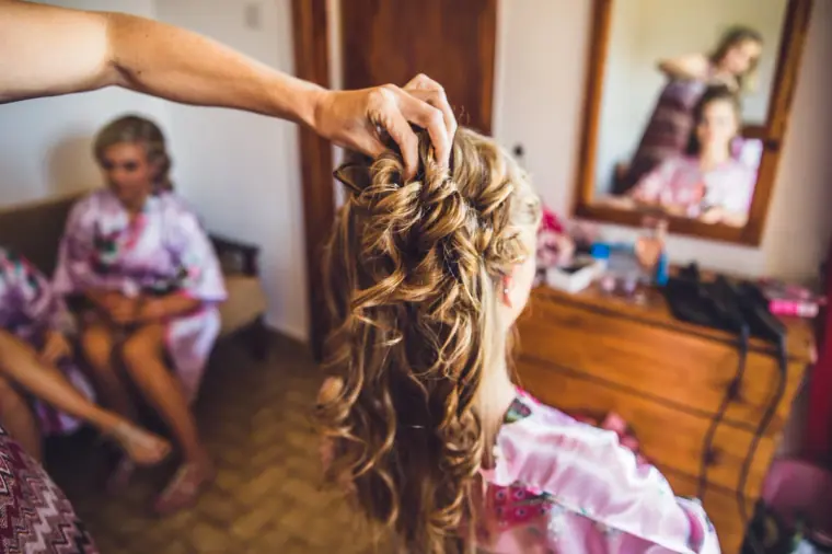 Bride's long curls being styled as a bridesmaid in a floral robe waits