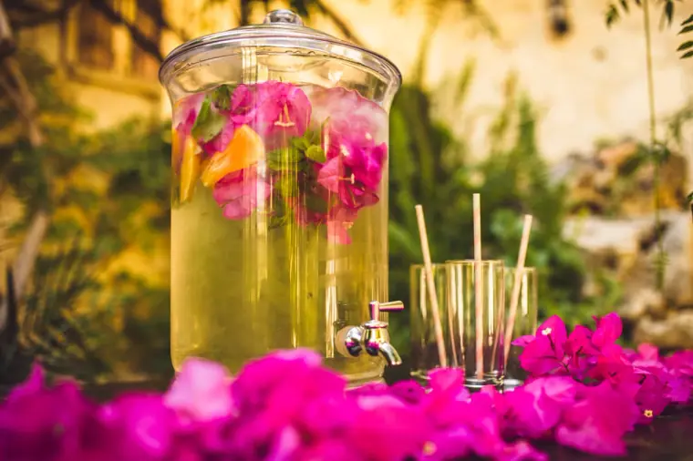 Glass drinks dispenser of lemonade with floating flowers on a bougainvillea-covered table